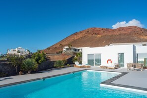 Un área de piscina moderna con una piscina azul cristalina, un edificio blanco y una montaña al fondo.