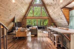 A spacious loft with a high ceiling, large windows offering a view of the forest, a cozy seating area, and a kitchen with a long counter.