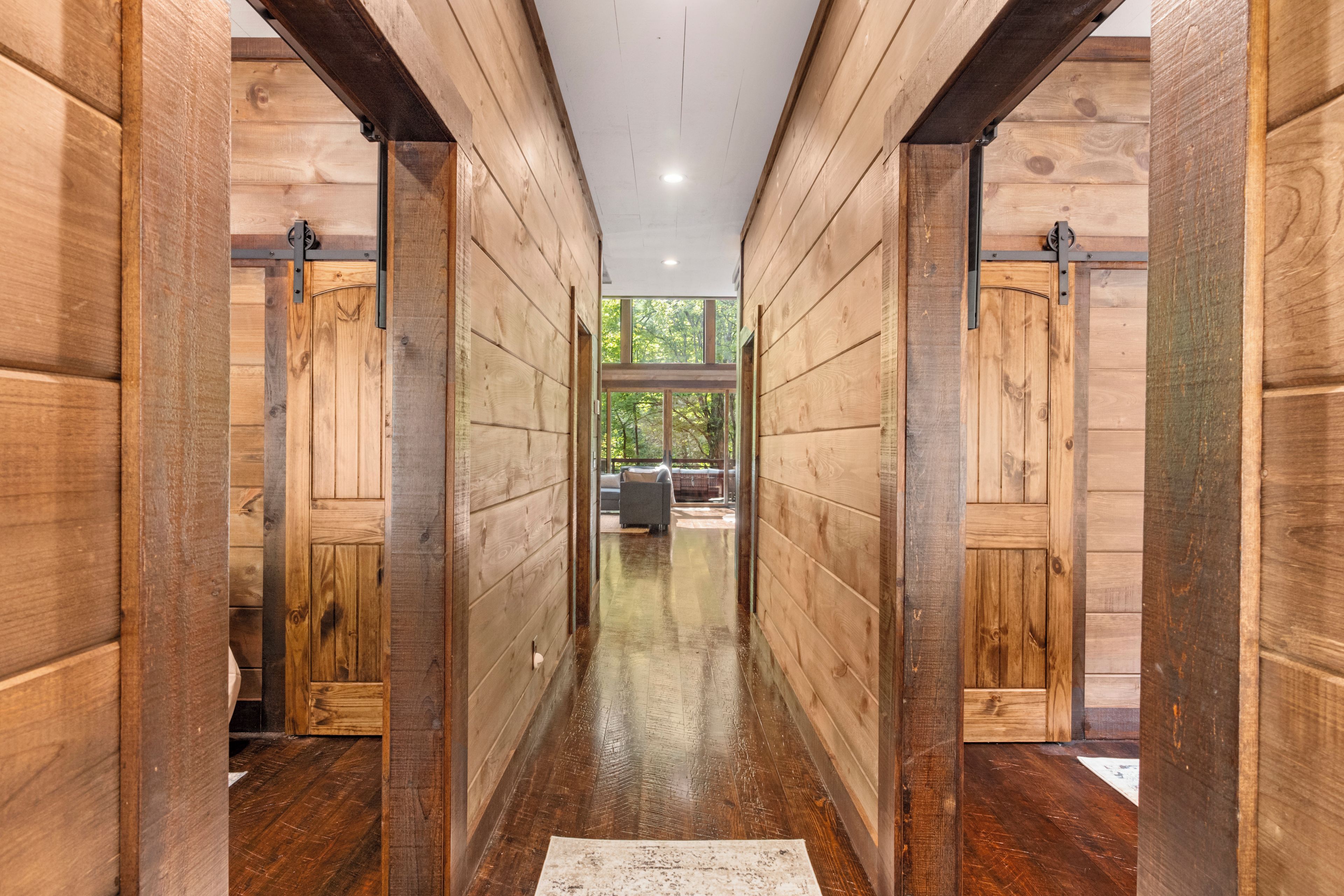 A wooden hallway with a view of a living area and a window overlooking greenery.
