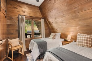 A wooden cabin room with two beds, a chair, a nightstand with a lamp, and a window with curtains.