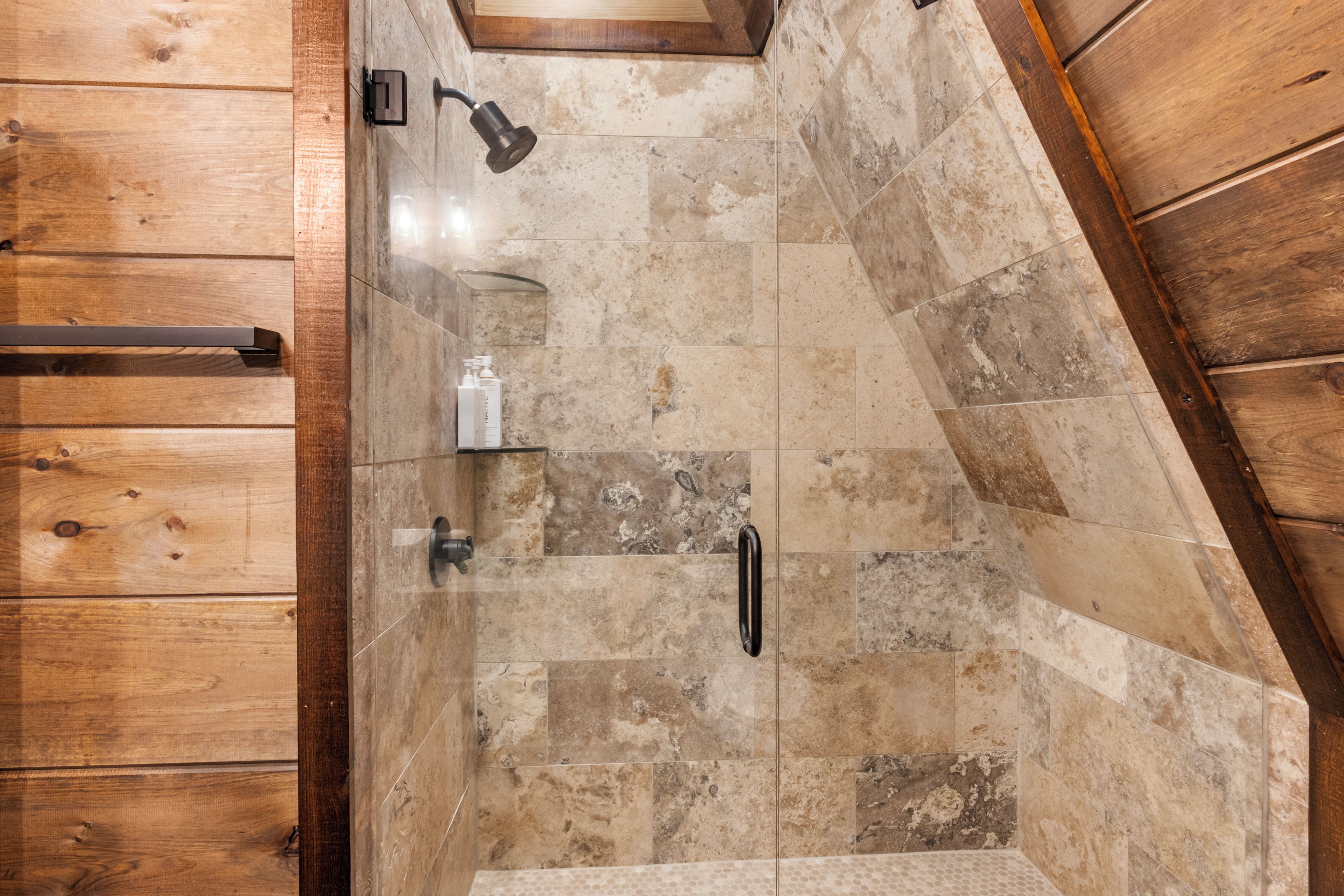 A shower area with a wooden door, a handheld showerhead, and a bottle of shampoo.