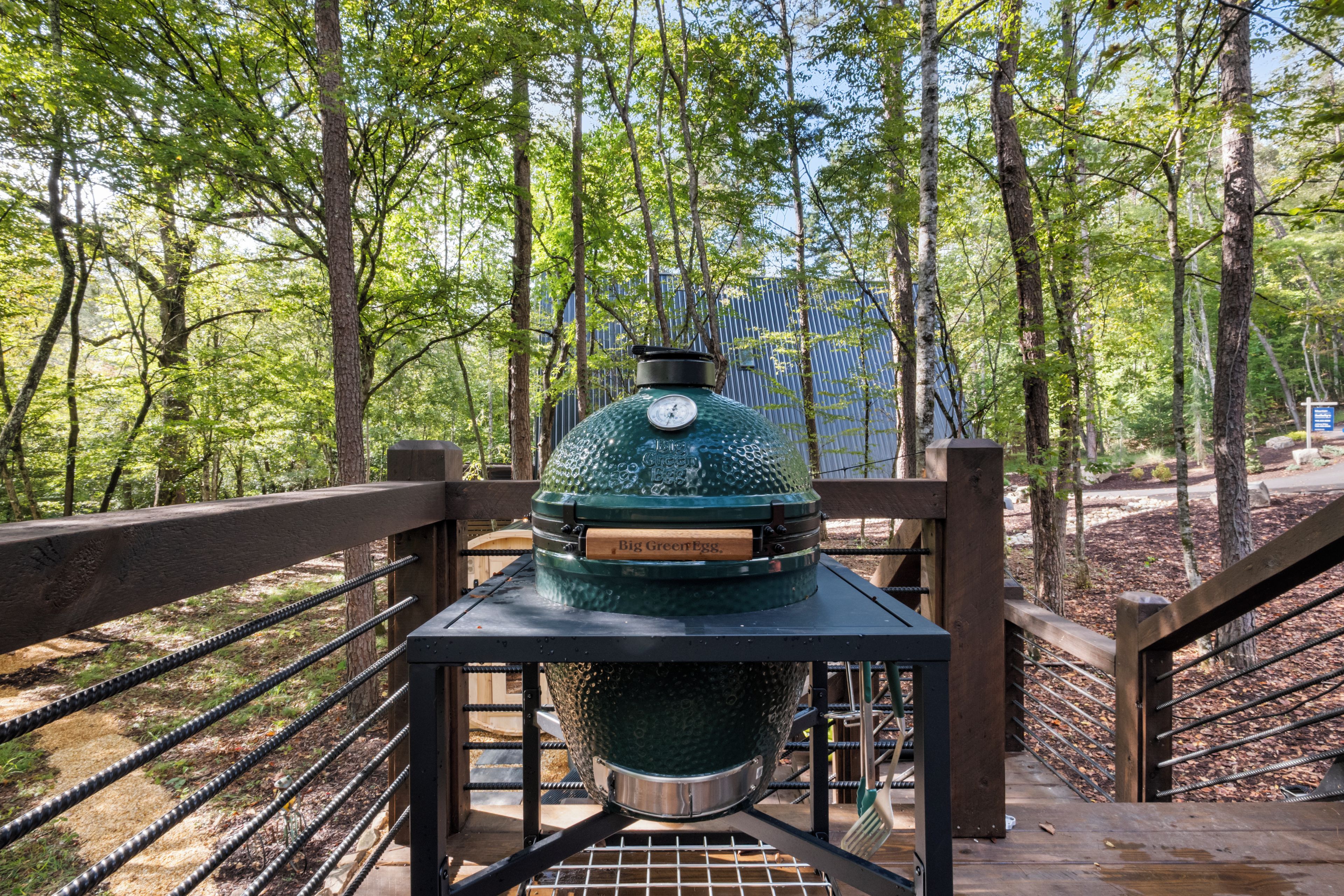 A green outdoor grill on a wooden platform with metal railings, surrounded by trees.