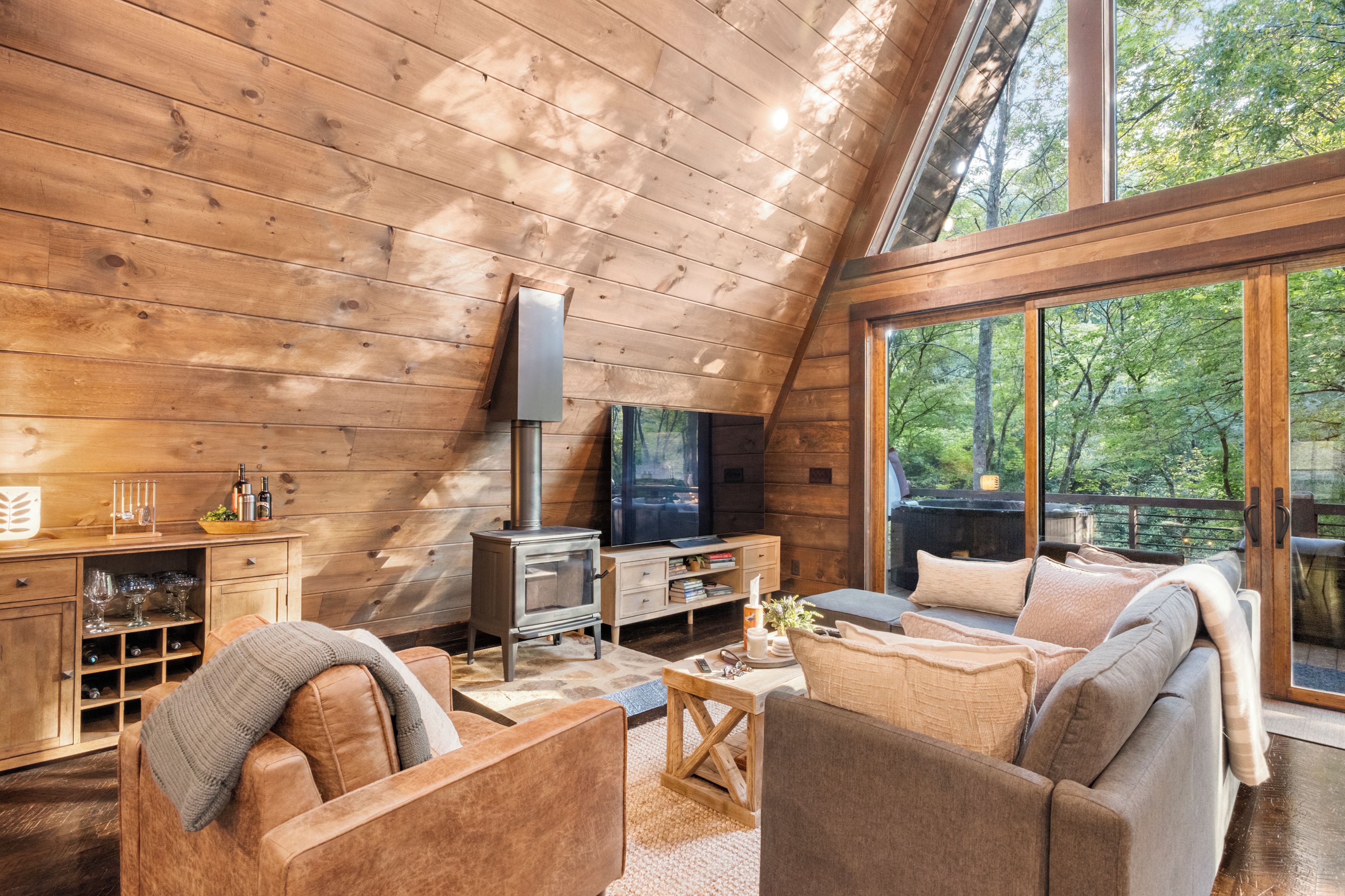 L’intérieur chaleureux d’un chalet, avec un décor en bois, une cheminée, un téléviseur et une grande fenêtre donnant sur la forêt.