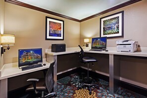 A hotel room with a desk, computer, printer, and two framed pictures on the wall.