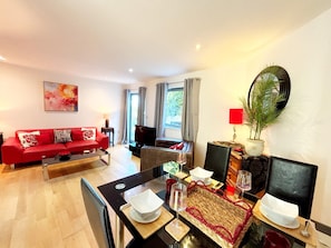 A modern living room with a red sofa, a dining table set for two, and a large window with curtains. - Two bedroom, two bathroom apartment with terrace close to Torquay seafront. (Torquay)