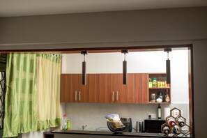 Une cuisine avec des meubles de rangement en bois, un luminaire suspendu, une fenêtre ornée d’un rideau vert et une étagère contenant des ustensiles de cuisine.