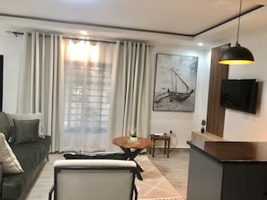 A modern living room with a sofa, a dining table, and a TV mounted on the wall. - Plush 1Bed Apt PHI Great East Rd (Lusaka)