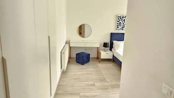 A narrow hallway leading to a bedroom with a white wardrobe, a round mirror, a small blue ottoman, and a bed with a blue headboard.