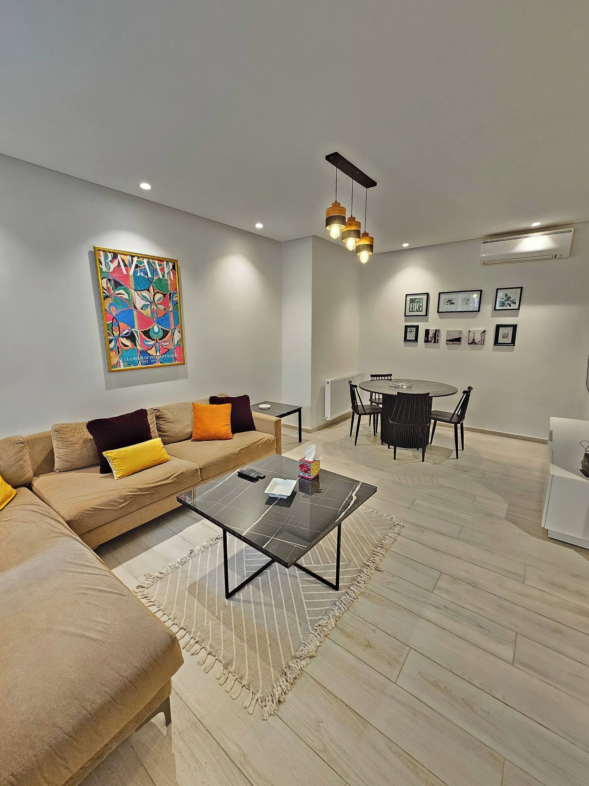 A modern living room with a sectional sofa, a glass coffee table, a dining area with a round table and chairs, and a large abstract painting on the wall.