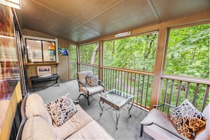 A covered porch with a sofa, chairs, and a table, overlooking a forested area.