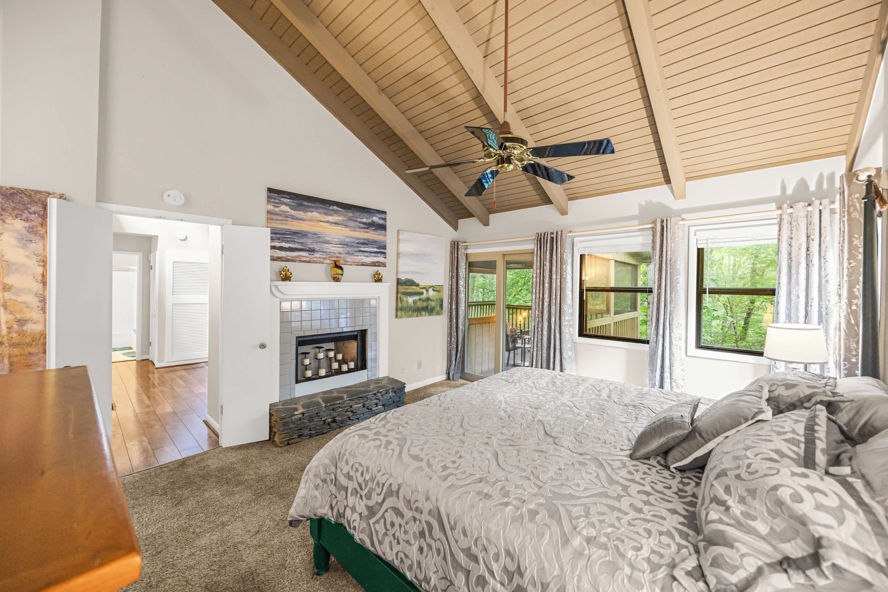 A bedroom with a large bed, a fireplace, a ceiling fan, and a view of the outdoors.