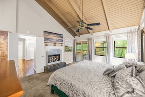 A bedroom with a large bed, a fireplace, a ceiling fan, and a view of the outdoors.