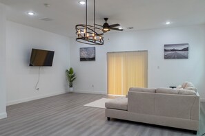 A modern living room with a flat-screen TV, a ceiling fan, and a large window with blinds. - Cherry Lux (Houston)