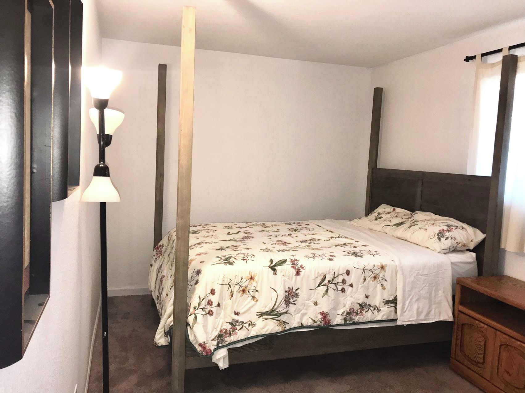A four-poster bed with a floral bedspread, a bedside table, and a floor lamp.