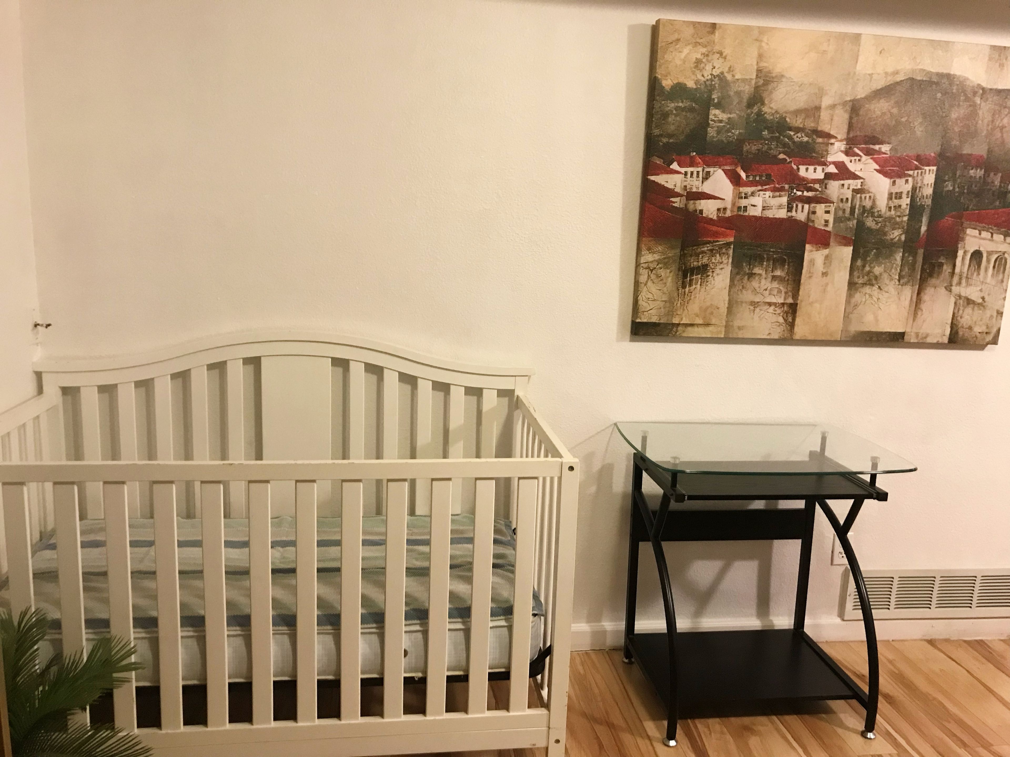 Una cuna blanca, una mesa con superficie de vidrio y un cuadro de un pueblo en la pared.