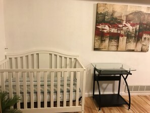 A white crib, a glass-top table, and a painting of a town on the wall.