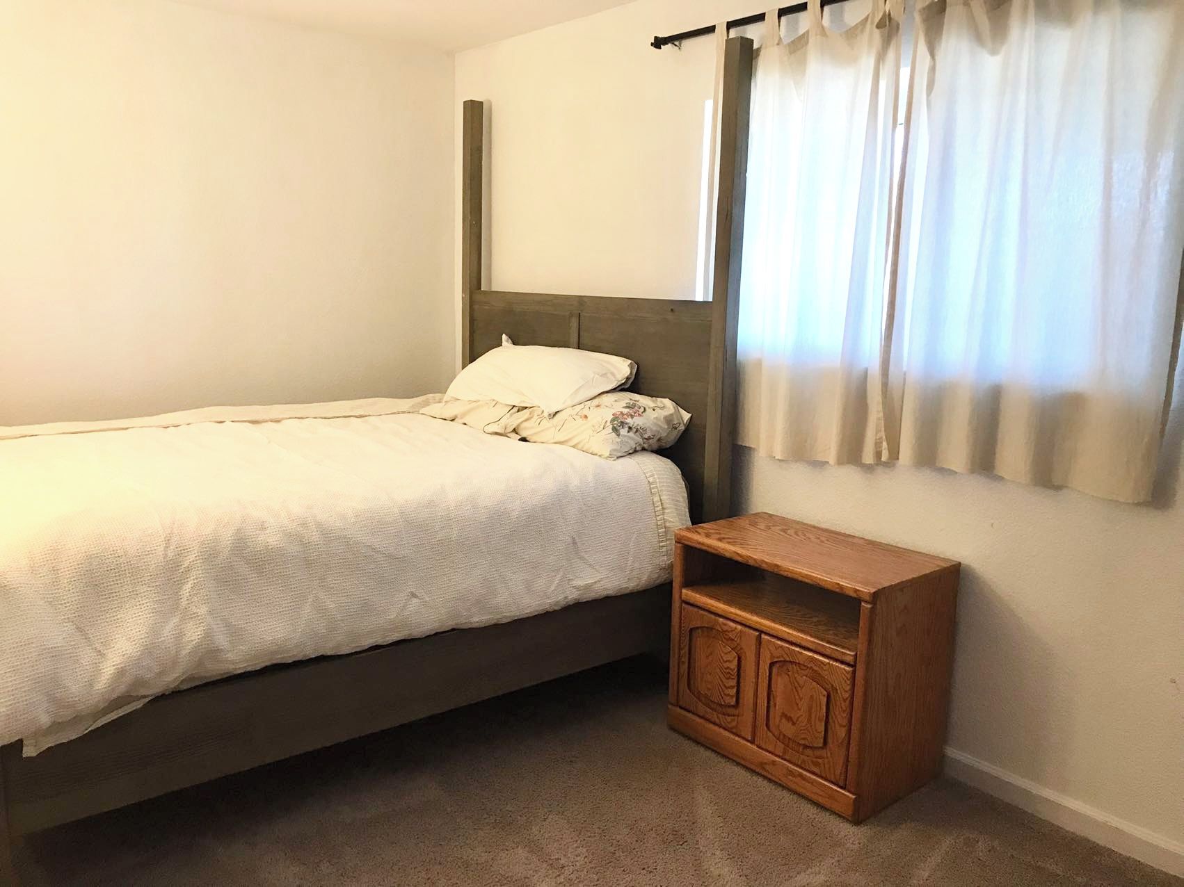 A single bed with a headboard, a wooden nightstand with drawers, and a window with curtains.