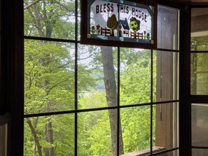 A window with a 'Bless This House' sign, a view of a forest, and a wooden structure.