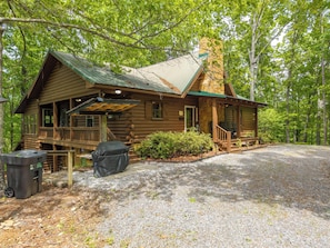 A log cabin with a porch, a covered grill, and a gravel driveway.