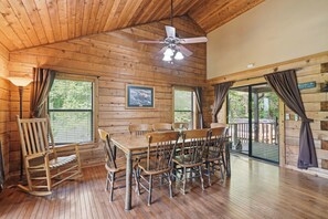 A wooden cabin interior with a dining table, chairs, a rocking chair, a ceiling fan, and large windows.