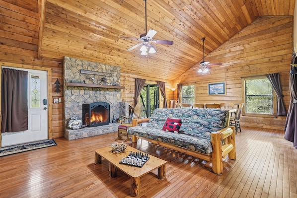 A cozy cabin interior with a stone fireplace, wooden furniture, and a chessboard on a coffee table.
