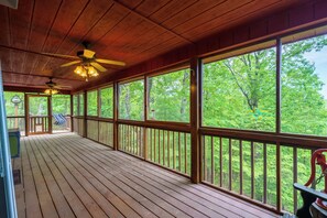A covered wooden deck with a ceiling fan, large windows, and a view of a forest. - Hanging Out in the Clouds (Blue Ridge)