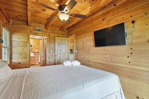 A wooden cabin bedroom with a bed, a ceiling fan, a TV mounted on the wall, and a door leading to a bathroom. - Hanging Out in the Clouds (Blue Ridge)