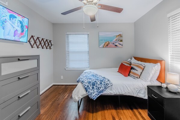 Un dormitorio con cama, cómoda, ventilador de techo, un cuadro y televisor en la pared.
