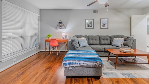 Een woonkamer met een grijze bank, een houten salontafel, een rode fauteuil, een blauw-witte gestreepte voetenbank en een plafondventilator, met een wandplank met decoratieve objecten.