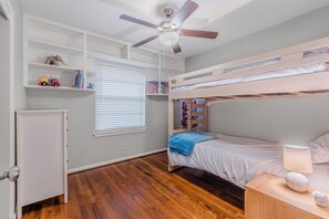 A bedroom with a bunk bed, a ceiling fan, a window with blinds, a shelf with toys, and a bedside table with a lamp. - Perfect 3-bedroom house w/ Smart TV, Patio, in peaceful Dallas neighborhood (Dallas)
