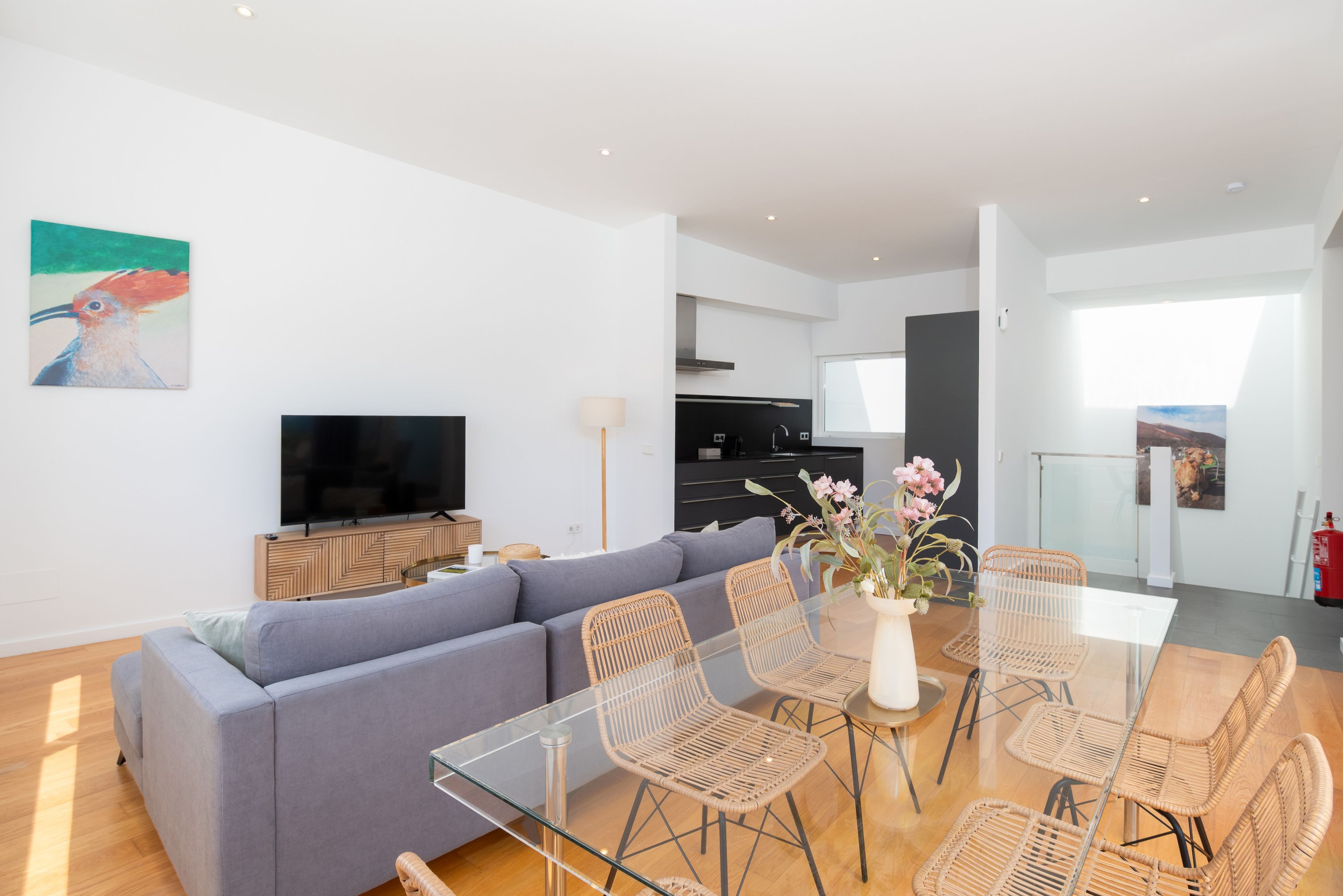 Una sala de estar moderna con sofá, mesa de comedor de vidrio, sillas de mimbre, televisor de pantalla plana y obra de arte en la pared.