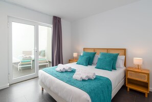 Ein Hotelzimmer mit einem großen Bett, einem Holzkopfteil, zwei Nachttischen mit Lampen, einer Schiebetür zum Balkon und Meerblick.