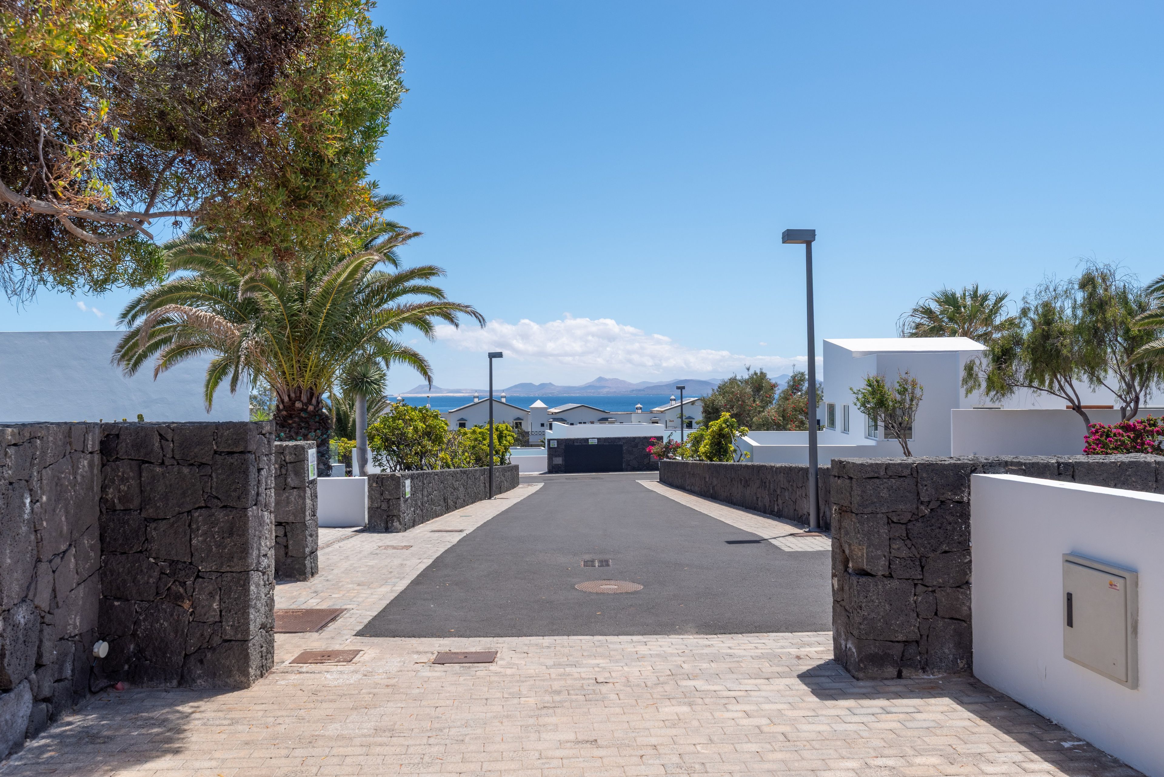 Un camino pavimentado que lleva a un edificio con muros de piedra y una vista despejada del mar y las montañas al fondo.