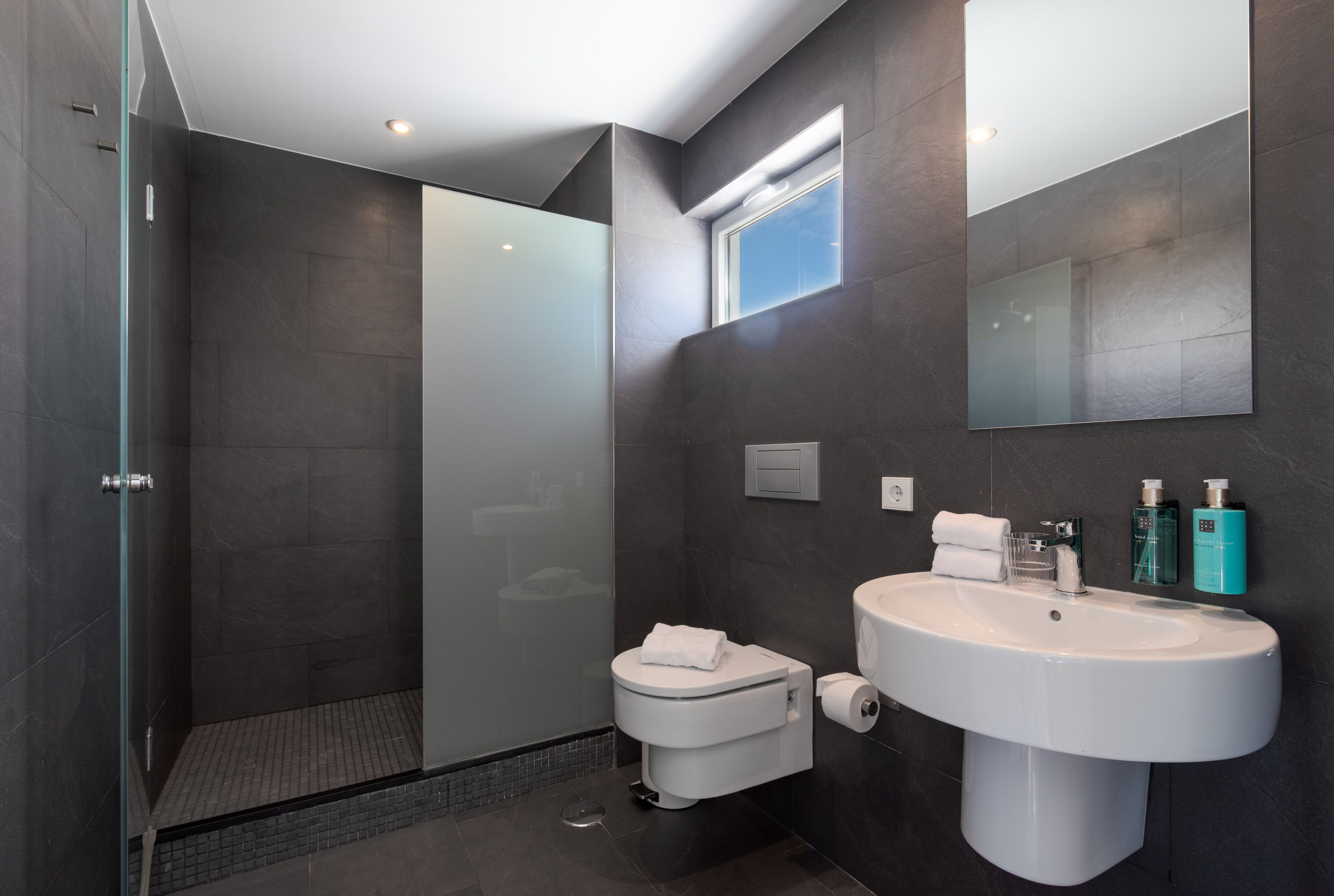 Un baño moderno con cabina de ducha de vidrio, inodoro, lavabo con espejo y artículos de aseo.