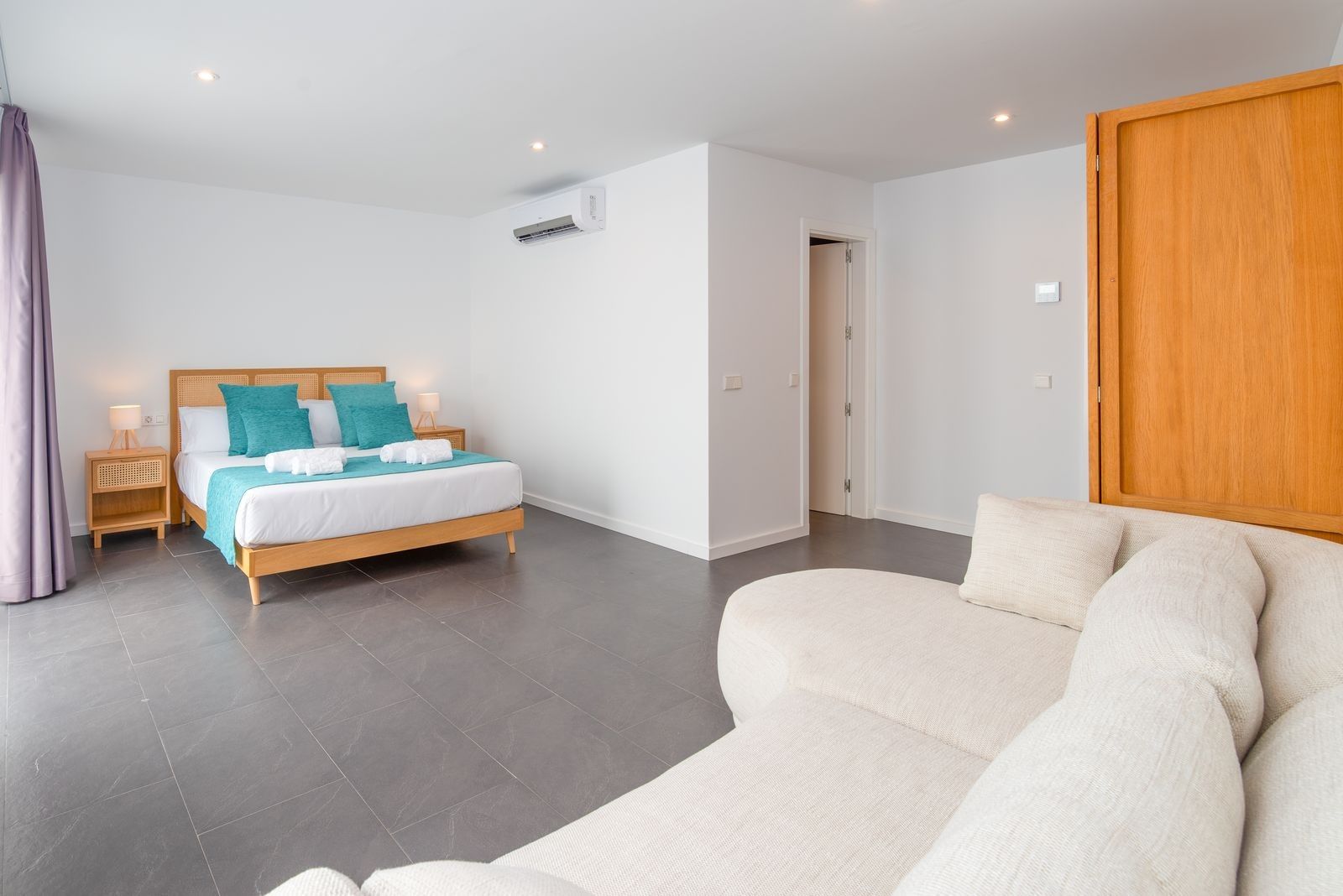 Ein modernes Hotelzimmer mit einem Bett, einem Sofa, einem Holzschrank und einer Tür zu einem weiteren Raum.