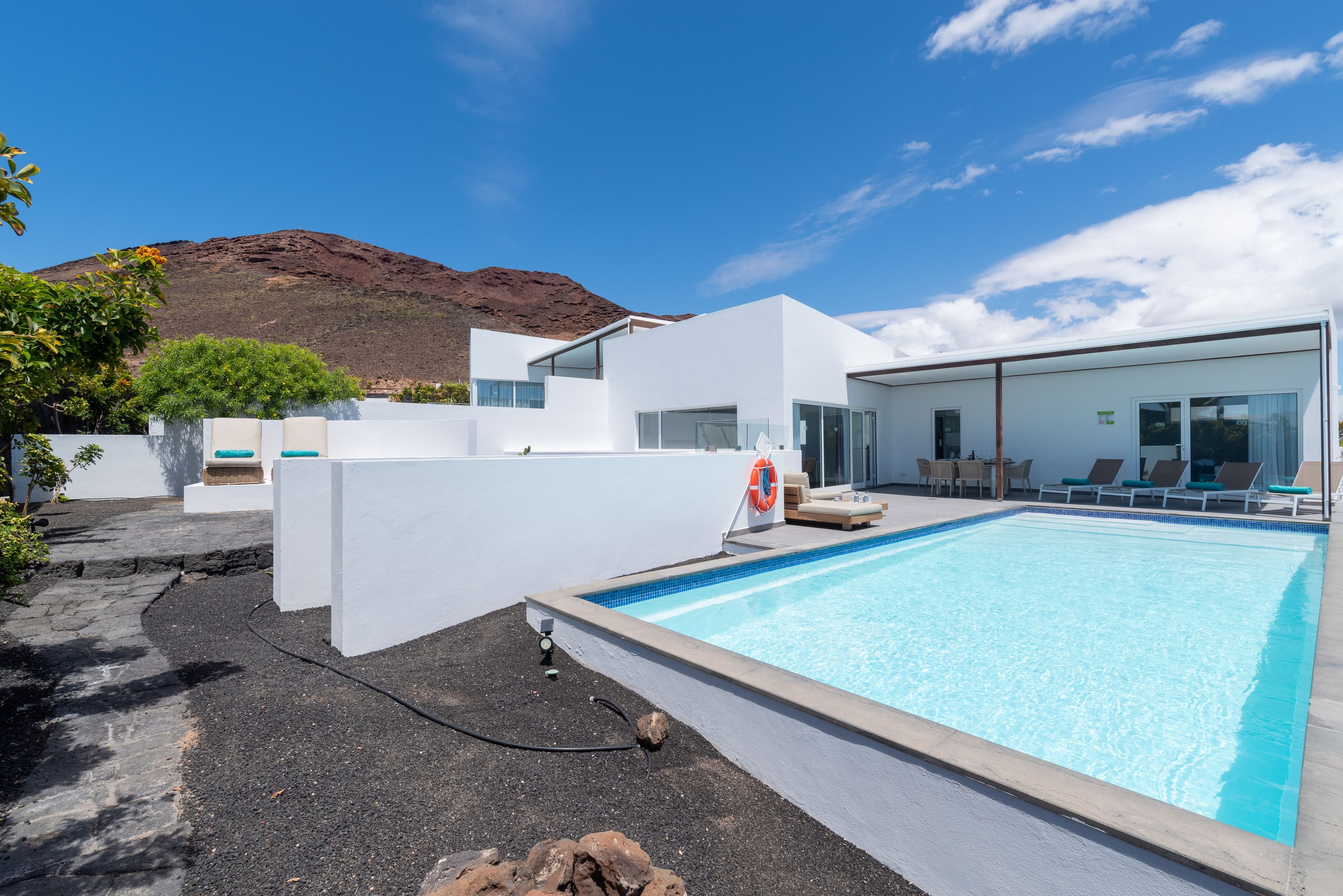 Un área de piscina moderna con una piscina azul cristalina, paredes blancas y una montaña al fondo.