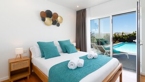 Une chambre d’hôtel moderne avec un grand lit, une literie de couleur turquoise, un élément décoratif mural, une table de chevet avec une lampe et une vue sur une piscine, visible à travers une porte-fenêtre coulissante.