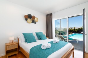 Quarto de hotel moderno com uma cama grande, roupa de cama verde-água, um painel decorativo na parede, mesa de cabeceira com abajur e vista para uma piscina através de uma porta de vidro deslizante.