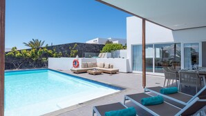 Un espace piscine extérieur moderne, avec des chaises longues et un canapé, un ciel bleu dégagé et un mur de pierre en arrière-plan.