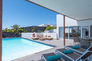 Uma área moderna com piscina ao ar livre, espreguiçadeiras e um sofá, céu azul límpido e uma parede de pedra ao fundo.