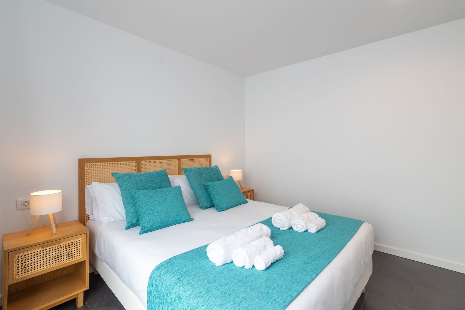 Una cama bien hecha con un edredón color turquesa, almohadas blancas y una manta turquesa a juego. Hay dos lámparas de noche sobre mesitas de madera.