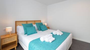Una cama bien hecha con un edredón color turquesa, almohadas blancas y una manta turquesa a juego. Hay dos lámparas de noche sobre mesitas de madera.