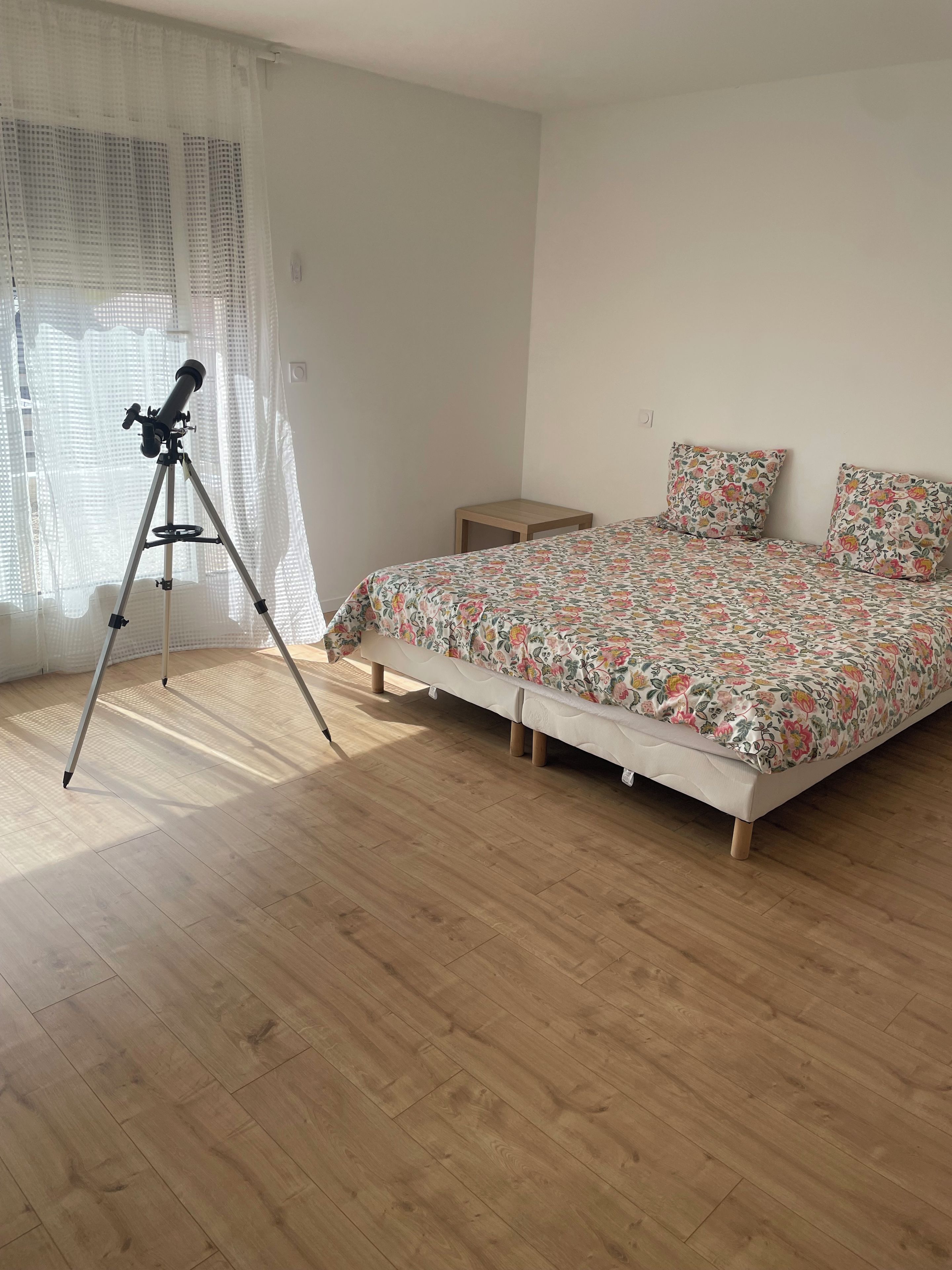Un dormitorio con piso de madera, una cama con ropa de cama floral, una mesita de noche pequeña y un telescopio.