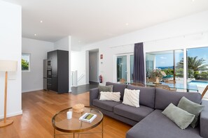 A modern living room with a grey sofa, a glass coffee table, and a view of a pool and palm trees. - Vacation Villa 'Ereza Royal Deluxe Hoopoe 104' with Mountain View, Private Pool and Wi-Fi (Playa Blanca)
