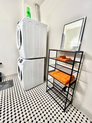 A laundry area with a stacked washer and dryer, a metal shelf with folded towels, and a black and white patterned floor. - Charming Vintage Loft in Bishop Arts (Dallas)