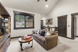 A living room with a sofa, coffee table, and a window overlooking a wooded area. - NEW! The Alders - Walk 2 Ski, Hot Tub, Fireplace (Dillon)