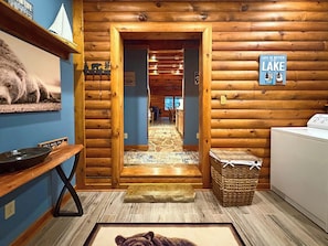 A cabin interior with a wooden wall, a welcome sign, a rug, a washing machine, and a view of the interior through a doorway.