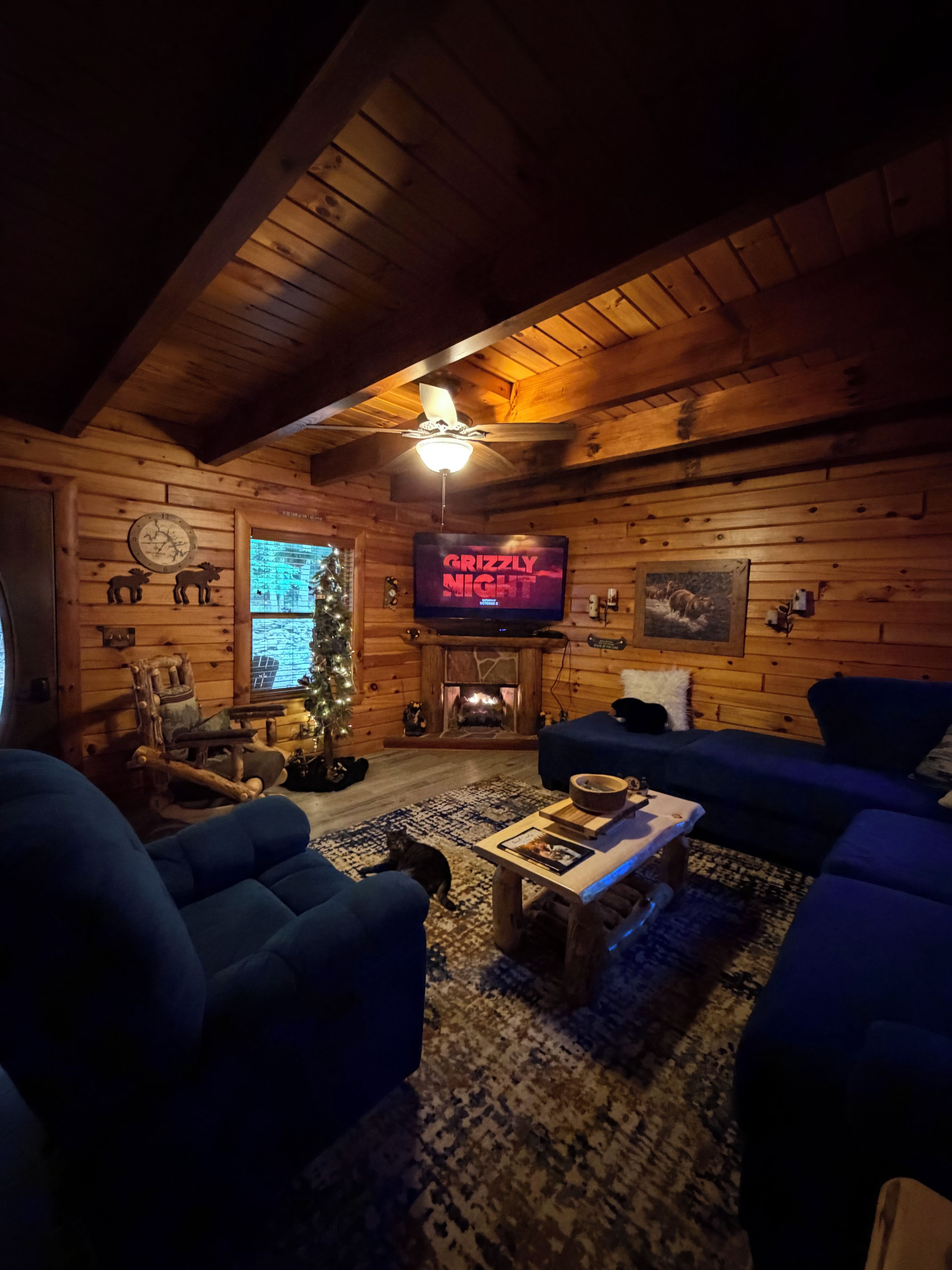 A cozy log cabin interior with a fireplace, a television displaying 'GRIZZLY NIGHT,' and a Christmas tree.