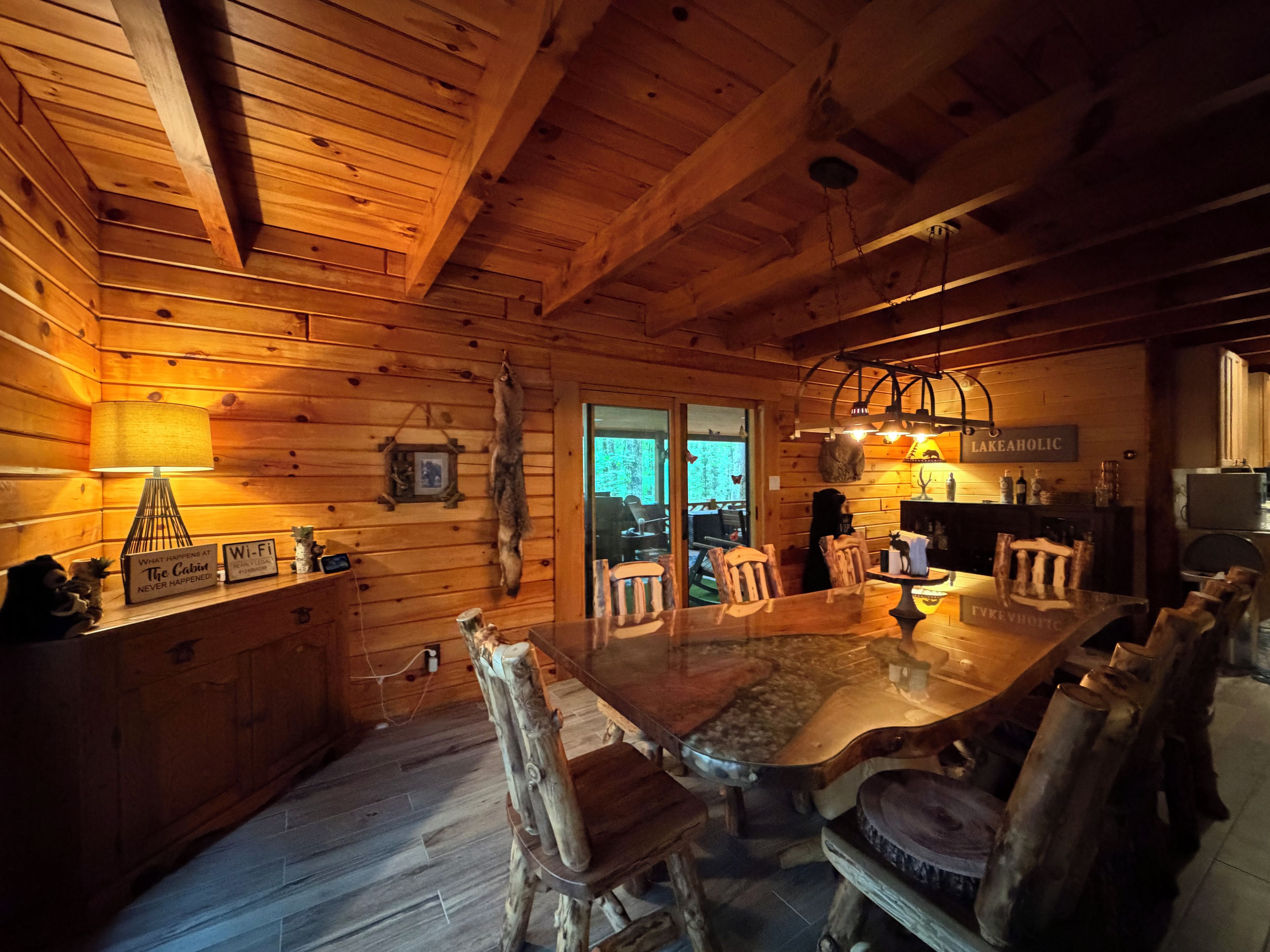 A wooden cabin interior with a large dining table, chairs, a hanging lamp, and a sign reading 'LAKEAHOLIC'.