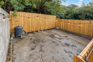 A wooden fence with string lights, a trash bin, and a concrete patio. - 3-bed Terrace House in Porth, Sleeps 6 (Porth)